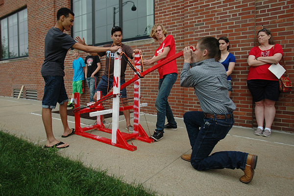 A team of four campers prepares to launch a tennis ball from a catapult device.